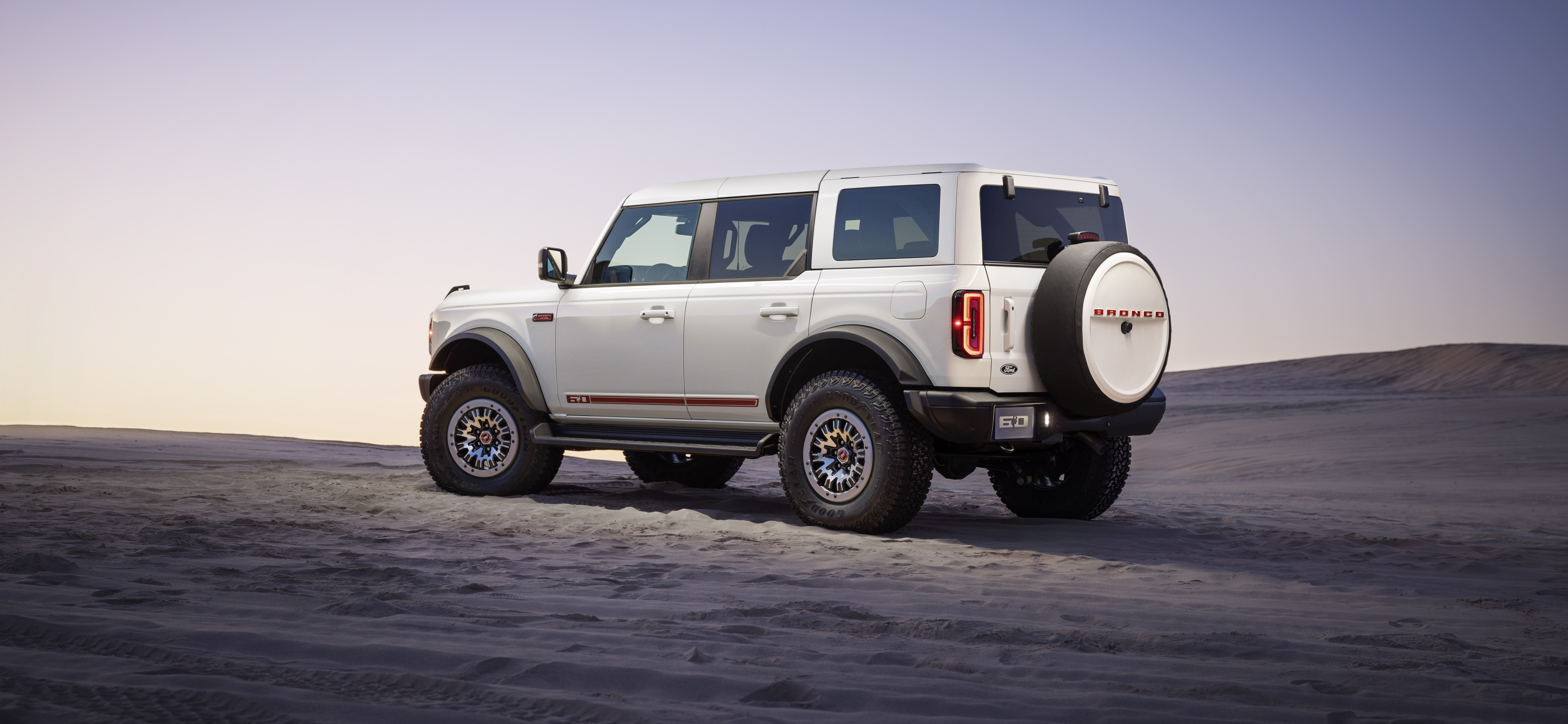 2026 Ford Bronco 60th Anniversary Package side view showing a white vehicle with red accents. Vehicle is on the sand with a purple and yellow dusk sky overhead.
